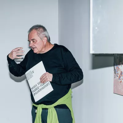 Marek Sobczyk stoi w przestrzeni wystawy, gestykuluje, w dłoni trzyma katalog do wystawy, z prawej strony widać fragment jego obrazu