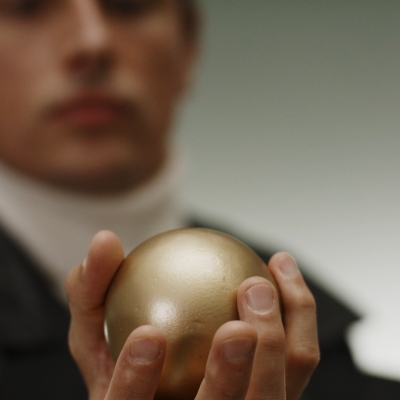 Jasmina Cibic, "The Gift", 2021, screenshot, courtesy of the artist: In the foreground, there is a golden ball held by a man. 