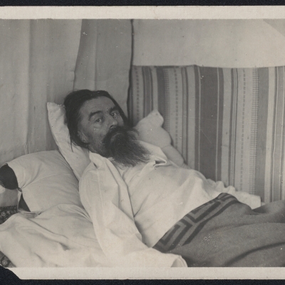 Autor nieznany, „Kazimierz Malewicz, leżący w łóżku”, 1934-35 r., fotografia czarno-biała, z kolekcji Muzeum Sztuki w Łodzi: Starszy mężczyzna z brodą i w białej koszuli, leży na łóżku przykryty kocem.