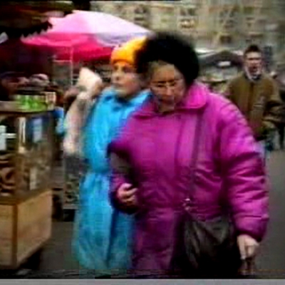 Alicja Żebrowska, Koniec mojego wieku, 1992-1995, VHS, dzięki uprzejmości artystki.: Niskiej jakości kadr wideo, z dwoma osobami na targowisku w jaskrawych kurtkach : fioletowej i turkusowej. 