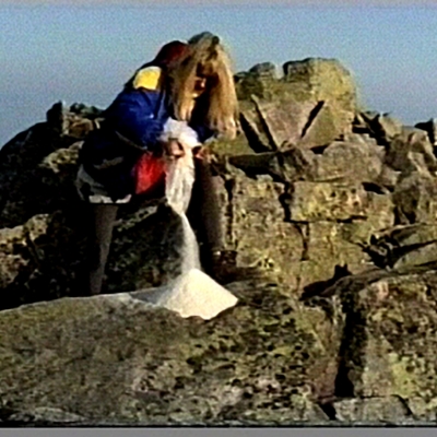 Alicja Żebrowska, Trans-fero, 1992, VHS, dzięki uprzejmości artystki: Artystka w sportowej kurtce wysypuje biały pył w szaro-brązowych skałach, w tle błękitne niebo.