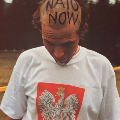 Piotr Wyrzykowski, NATO Now!, 1994, fotografie, dzięki uprzejmości artysty: Zdjęcie mężczyzny w białej koszulce z godłem polski. Mężczyzna pochyla częściowo zgoloną głowę z napisem Nato Now.