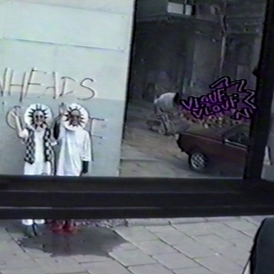 Wspólnota Leeeżeć, Pozdrowienie, 1990, happening, zapis na taśmie VHS, dzięki uprzejmości Andrzeja Miastkowskiego: Niskiej rozdzielczości kadr z wideo, przez szybę autobusu widać dwie machające postaci w białych ubraniach, mają  promieniste - przypominające słoneczko nakrycia głowy.