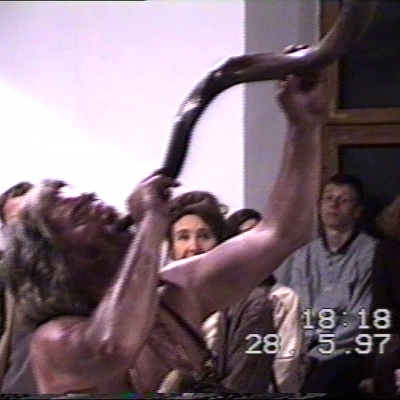 Zbigniew Warpechowski, New Age - Róg pamięci, 1997, kadr z filmu, VHS, dzięki uprzejmości Galerii Monopol: Niskiej rozdzielczości kadr z wideo, mężczyzna w długich siwych włosach dmie w róg, jego ciało wysmarowane jest na brązowo. 