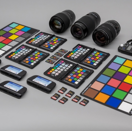 Widok z góry na kompletny zestaw sprzętu fotograficznego i do kalibracji barw ułożony na jednolicie szarym podłożu. W skład zestawu wechodzą obiektywy, tablice ColorChecker, karty pamięci, czytniki oraz światłomierz.