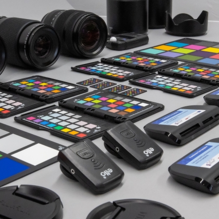 Zdjęcie ukazujące z bliska profesjonalne akcesoria fotograficzne ułożone na szarym tle. W kadrowanej przestrzeni widać czarne obiektywy, wzorce barwne (ColorChecker) z kolorowymi kwadratami oraz drobne urządzenia elektroniczne i czytniki kart.
