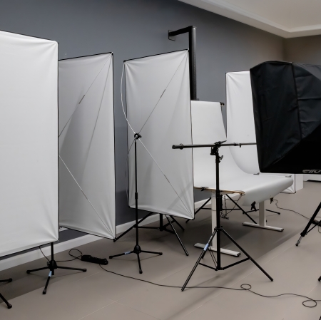 Przedstawia profesjonalne studio fotograficzne z ciemnoszarymi ścianami i jasną podłogą. W pomieszczeniu rozstawiono sprzęt oświetleniowy, w tym wysokie białe blendy dyfuzyjne na statywach oraz czarny softbox skierowany w stronę stanowiska fotograficznego.