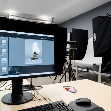Widok zza stanowiska komputerowego w studio fotograficznym, gdzie na dużym, profesjonalnym monitorze z osłoną wyświetlane jest oprogramowanie do obróbki zdjęć, pokazujące miniatury i powiększenie białego przedmiotu.[cite: 1] W tle, po prawej stronie, widać rozstawiony sprzęt studyjny, w tym białe tło fotograficzne oraz czarny softbox oświetleniowy marki Elinchrom na statywie.
