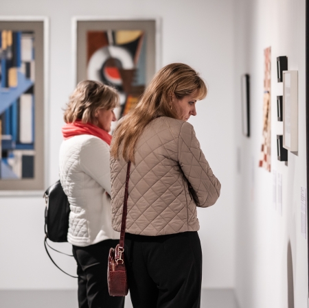Dwie kobiety stojące blisko ściany w galerii sztuki i z zaciekawieniem studiujące wiszące przed nimi abstrakcyjne dzieła.