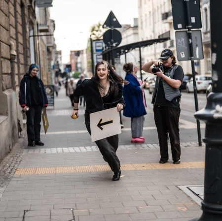 Dynamiczne ujęcie na miejskim chodniku, gdzie uśmiechnięta dziewczyna w ciemnym ubraniu biegnie przed siebie, trzymając w ręku białą planszę z czarną strzałką w lewo. W tle i po bokach przyglądają się jej znajomi, w tym jedna osoba robiąca zdjęcie profesjonalnym aparatem.