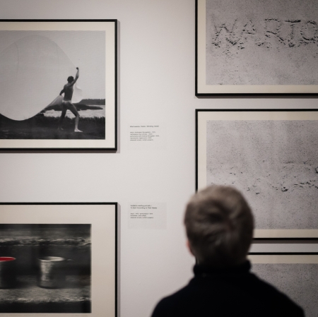 Odwrócona tyłem osoba przygląda się czterem czarno-białym, oprawionym w ramy fotografiom na białej ścianie galerii. Jedno ze zdjęć przedstawia naczynia z czerwonym akcentem, inne – napisy na piasku.