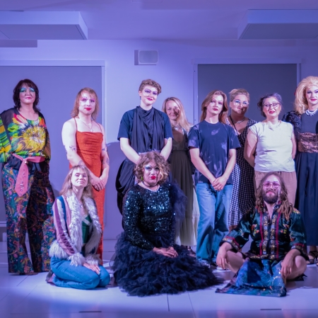 Grupowe zdjęcie dziesięciu osób w różnorodnych, artystycznych i dragowych stylizacjach, pozujących razem na jasnej scenie w chłodnym, niebieskawym oświetleniu – część z nich siedzi z przodu, a reszta stoi z tyłu.