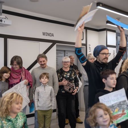 W nowoczesnym korytarzu z napisami na ścianach stoi uśmiechnięta grupa dorosłych i dzieci, wśród których wyróżnia się mężczyzna w okularach radośnie unoszący w górę kolorowe kartki.