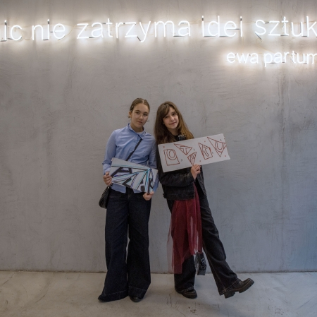 Dwie nastolatki stoją obok siebie na tle szarej ściany z neonowym napisem i z uśmiechem prezentują do zdjęcia swoje podłużne, artystycznie ozdobione kartony.
