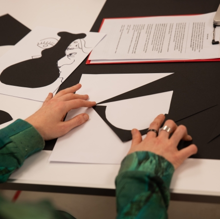Widok z góry na stół warsztatowy, na którym leżą gotowe, czarne wycinanki naklejone na biały papier, przybory do pisania, pojemnik z nożyczkami oraz kubek z herbatą. W tle widoczne są dłonie jednej z uczestniczek.