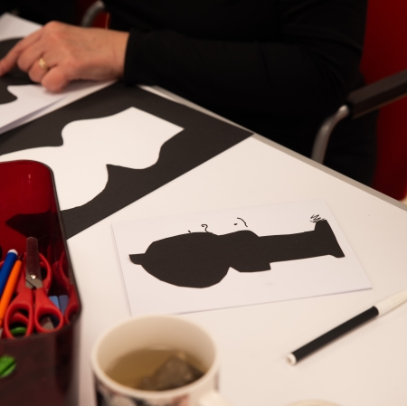 Zbliżenie z góry na dłonie osoby w zielonym ubraniu, która układa na stole wycięte, czarno-białe elementy geometryczne. Obok leżą arkusze papieru z grafikami oraz schowany w podkładce dokument.