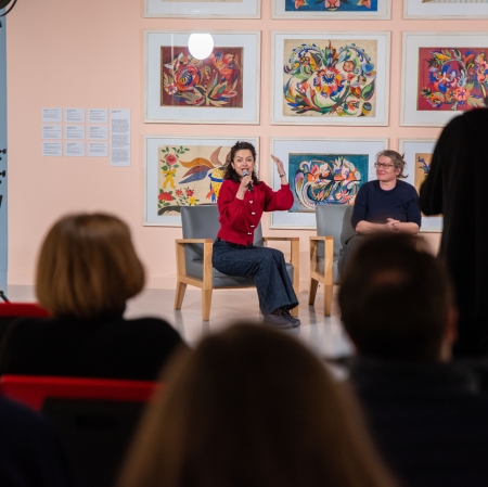Kobieta w czerwonej bluzce siedzi na scenie w fotelu i mówi do mikrofonu, gestykulując uniesioną dłonią w stronę publiczności. Spotkanie odbywa się na tle różowej ściany z galerią obrazów, a z boku widać sylwetkę operatora filmującego wydarzenie