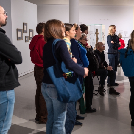 Zdjęcie przedstawia grupę ludzi zwiedzających wystawę w nowocześnie urządzonej galerii sztuki lub muzeum. Zgromadzeni stoją tyłem lub profilem do obiektywu, z uwagą oglądając eksponaty, obrazy oraz tablice informacyjne umieszczone na białych ścianach.
