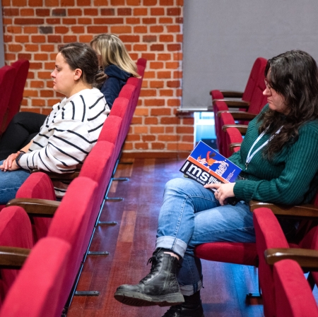 Trzy kobiety siedzą w rzędach czerwonych foteli kinowych; jedna z nich, w zielonym swetrze i ciemnych butach, trzyma na kolanach kolorowy folder z napisem "MS Muzeum Sztuki". Sala ma drewniany parkiet, szare wykończenie ścian oraz fragment z czerwonej cegły w tle.