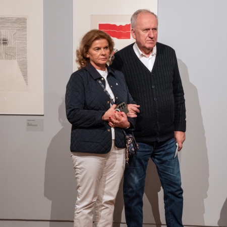 Starsze małżeństwo stoi blisko siebie w galerii sztuki na tle dwóch nowoczesnych, oprawionych grafik. Kobieta w pikowanej, granatowej kurtce i białych spodniach trzyma telefon, a stojący obok niej mężczyzna w ciemnym kardiganie trzyma ją pod rękę.