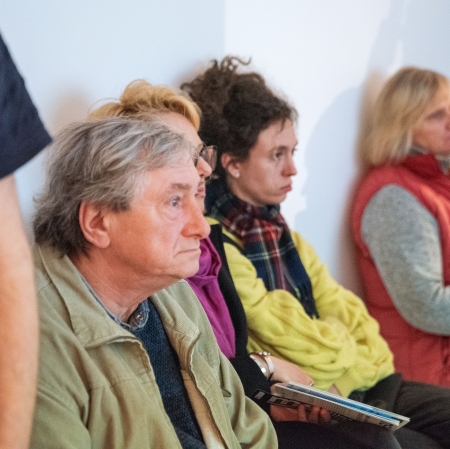 Ujęcie przedstawiające rząd osób siedzących blisko siebie pod ścianą i uważnie patrzących w jednym kierunku poza kadr. Na pierwszym planie siedzi mężczyzna w jasnej, rozpiętej kurtce, a obok niego widoczne są kolejne osoby, w tym kobieta w jaskrawożółtym swetrze i szaliku w kratę.