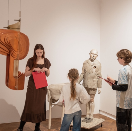 Dwoje dzieci z podkładkami do pisania stoi przed rzeźbą stojącego chłopca w muzeum, a towarzysząca im młoda kobieta w brązowej sukience trzyma czerwony arkusz papieru. Po lewej stronie kadru, na pierwszym planie, wisi nowoczesna, warstwowa rzeźba z drewna lub pleksi.
