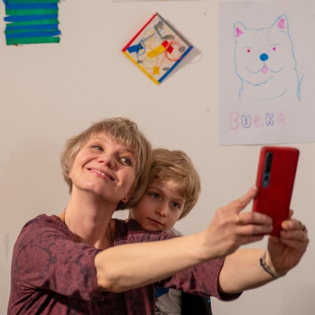 Pionowy portret uśmiechniętej kobiety z krótkimi, blond włosami oraz małego chłopca o jasnych włosach, który wygląda zza jej ramienia. Kobieta trzyma w wyciągniętej dłoni czerwony smartfon, robiąc im wspólne zdjęcie na tle ściany z dziecięcymi rysunkami.