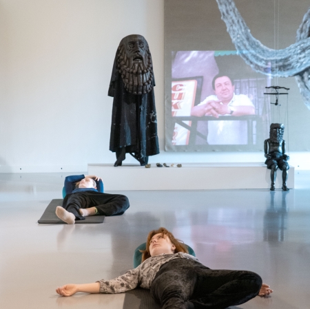 Perspektywa z głębi galerii sztuki, gdzie na pierwszym planie dwie osoby leżą tyłem na matach rozłożonych na błyszczącej podłodze. W tle widać biały podest z rzeźbami przedstawiającymi postacie ludzkie oraz duży ekran z projekcją wideo.