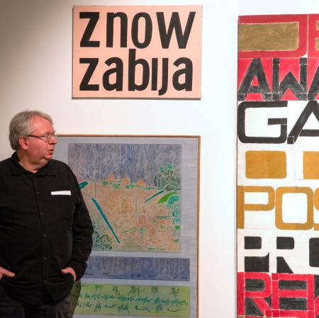 Mężczyzna w okularach i czarnej koszuli stoi w galerii sztuki na tle nowoczesnych obrazów, z których jeden przedstawia duży, czarny napis „znów zabija” na różowym tle.