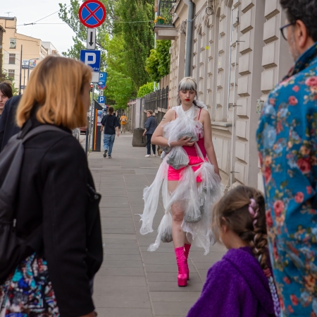 Dziewczyna w awangardowej kreacji z folii i z różowymi elementami stroju idzie chodnikiem wzdłuż zabytkowej kamienicy, przyciągając wzrok przechodniów.