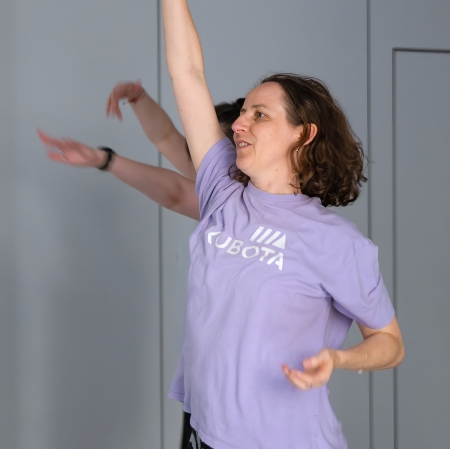 Kobieta o ciemnych, falowanych włosach, ubrana w fioletowy t-shirt i wzorzyste legginsy, uśmiecha się, wykonując taneczny gest z wysoko uniesioną prawą ręką na tle szarej ściany.