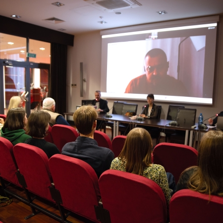 Zdjęcie przedstawia widok z tyłu na publiczność siedzącą w czerwonych fotelach sali konferencyjnej, która uczestniczy w spotkaniu z prelegentami oraz mężczyzną wyświetlanym na dużym ekranie.
