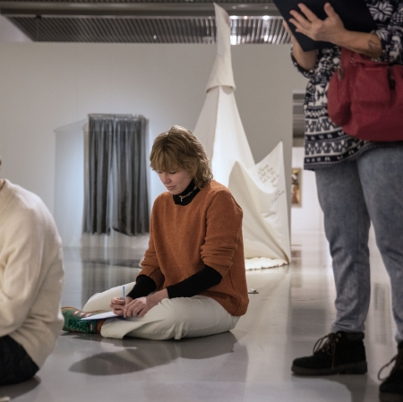 Trzy osoby znajdują się w jasnej, nowoczesnej galerii sztuki, gdzie dwie z nich siedzą na podłodze, rysując w notesach. W tle widać białą instalację przypominającą namiot oraz szare zasłony.