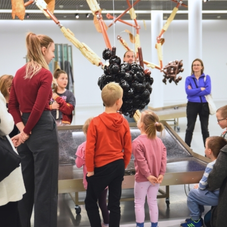 a group of adults and children in the exhibition space listening to an educator showing around the exhibition 
