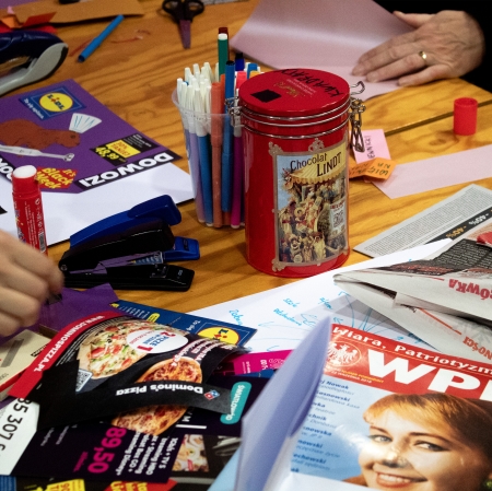Stół z rozłożonymi materiałami do tworzenia kolaży, w tym wycinkami z gazet i ulotek, kolorowymi papierami, pisakami, klejem, zszywaczami i czerwoną metalową puszką. Widoczne ręce dwóch osób pracujących nad projektem.