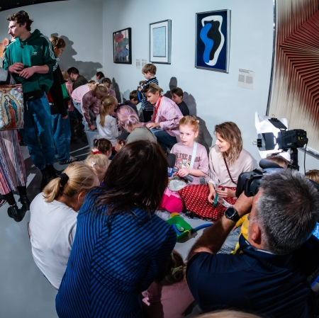 Kadr uchwycony szerokim kątem pokazuje oświetloną lampą błyskową grupę warsztatową na podłodze galerii, rejestrowaną przez profesjonalnego operatora kamery.