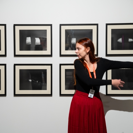Kobieta z identyfikatorem personelu, ubrana w czarną bluzkę i czerwoną spódnicę, dynamicznym gestem rozłożonych rąk wskazuje na serię czarno-białych fotografii wiszących na ścianie.