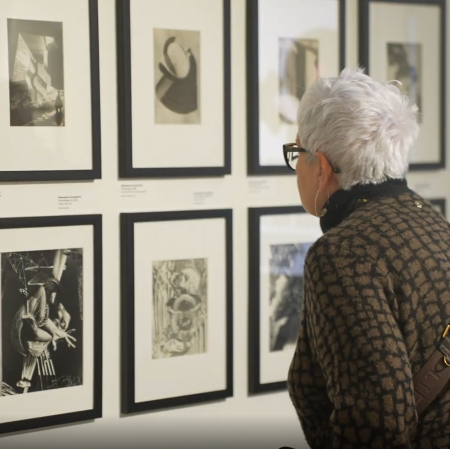Starsza osoba z krótkimi siwymi włosami ogląda z bliska czarno-białe fotografie oprawione w ramy na ścianie galerii.