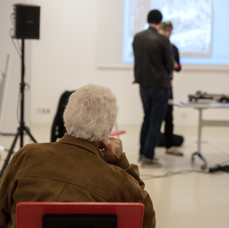 In the foreground, a man sits backward. In the background, people talking and a screen are visible.