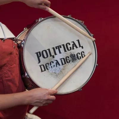  The photo shows a white snare drum with the writing "Political decadence" on it, the photo is taken from above. A person wearing a red overall is holding a snare drum and drumsticks, the background is also red.