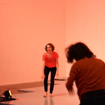 Młoda kobieta w pomieszczeniu o ciepłej, różowej barwie światła, wykonuje ruch teneczny, na pierwszym planie kilka osób stojących tyłem do obiektywu