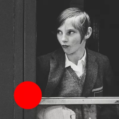 A black-and-white photograph presents a young girl on the train. She is looking outside small train window. There is a big, orange dot in the bottom left corner. 
