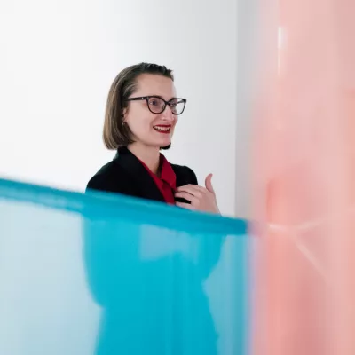 Młoda kobieta stoi w przestrzeni wystawy, mówi coś i gestyluje, jej dolną część ciała przesłania fragment instalacji w postaci półtransparentnej tkaniny w kolorze niebieskim i różowym