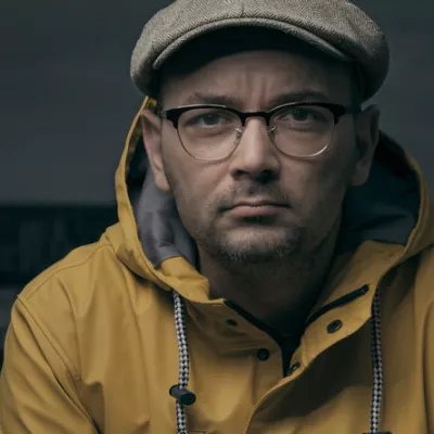 Mężczyzna w okularach, kaszkiecie i żółtej kurtce patrzy w obiektyw.