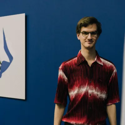Młody mężczyzna o blond włosach, w okularach, ubrany w czerwoną koszulę w białe poziome przetarcia, stoi na tle niebieskiej ściany, na której wisi wydruk komputerowy w kolorach: białym, czarnym i niebieskim