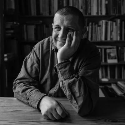 Czarno-białe zdjęcie. Mężczyzna siedzący na tle biblioteczki z książkami, podpiera jedną ręką głowę, druga jego ręka spoczywa na stole. Mężczyzna uśmiecha się. 