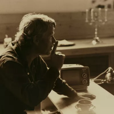 The man is sitting at the desk. He is putting his finger to lips.