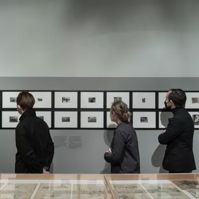 Na pierwszym planie gablota z rysunkami pod szkłem, trzy osoby w ciemnych ubraniach oglądają rzędy zawieszonych na szarej ścianie małych fotografii, oprawionych w czarne ramki.