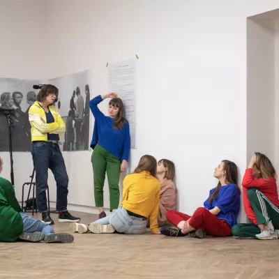 There are seven women in an exhibition hall. They are dressed in colorful clothes. Five of them are sitting on the floor, two are standing. Behind them, there is a camera on a tripod.