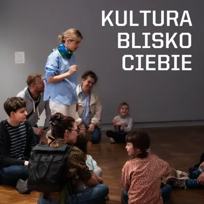 Grupa dorosłych i dzieci siedzi na podłodze w przestrzeni muzealnej podczas zajęć. Na grafice widoczny jest napis „KULTURA BLISKO CIEBIE”.  